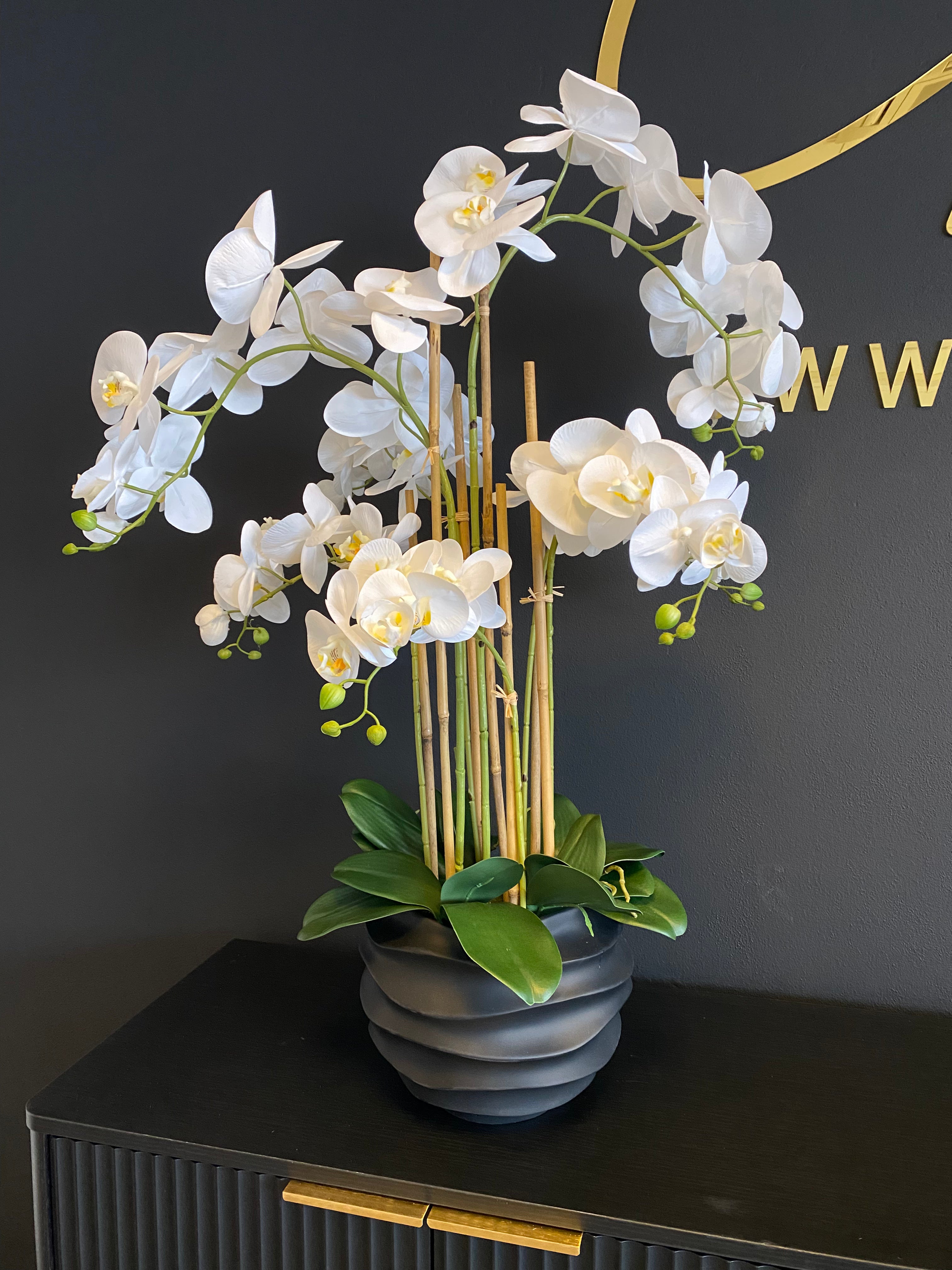 Witte Real Touch kunstorchidee van 85 cm met meerdere bloemtakken, groene bladeren en een donkergrijze geribbelde pot.